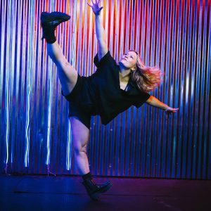a dancer in black shorts and t-shirt with one leg and arm raised in an active pose and pink and blue LED lights in the background