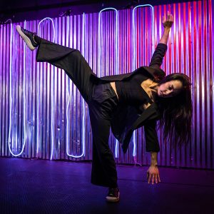 a dancer in black pants, shirt and jacket strikes an active pose with right arm and leg lifted and pink and purple LED lights in the background