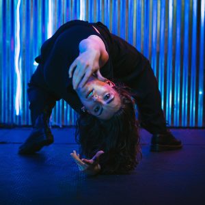 a dancer in black pants and shirt bends backwards toward the camera with blue LED lights in the background