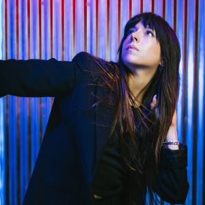A dancer in a black shirt and blazer with long dark hair looks up and to the left, with pink and blue LED lights in the background