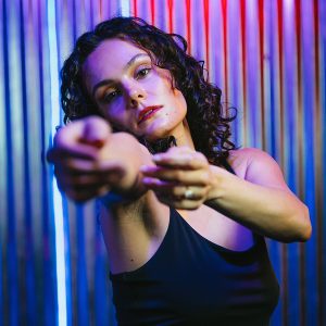 a dancer in a black tank top pictured from the waist up reaches toward the camera with pink and blue LED lights in the background