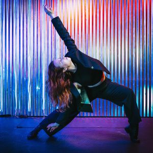 a dancer in black pants, shirt and jacket strikes a lunging pose with pink and blue LED lights in the background